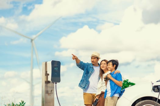 Concept of progressive happy family enjoying their time at wind farm with electric vehicle. Electric vehicle driven by clean renewable energy from wind turbine generator for charging station.