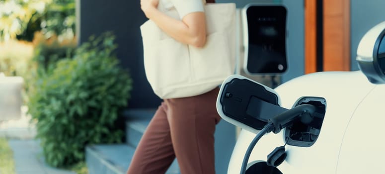 Focus image of electric vehicle recharging battery at home charging station with blurred woman walking in the background. Progressive concept of green energy technology applied in daily lifestyle.