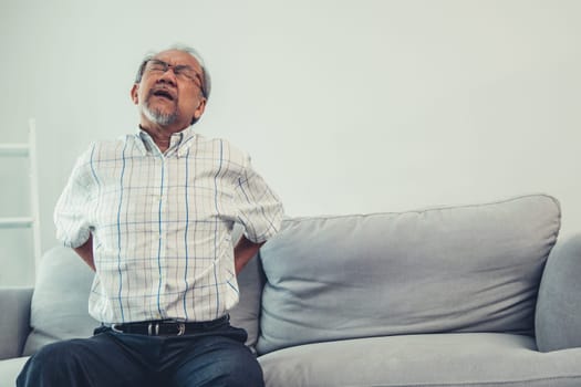 An agonizing senior man in need of assistance while sitting on his sofa at home, suffering from back pain. Senior care, nursing home for pensioners, deteriorating health of old age.