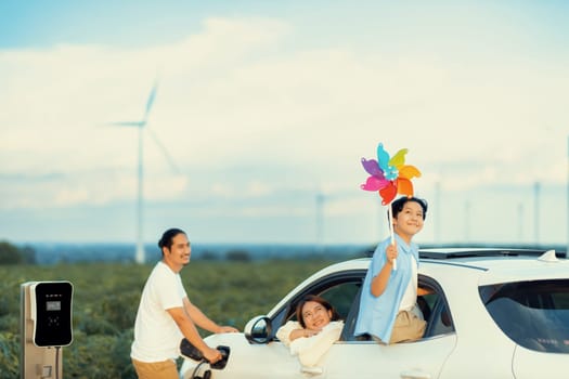 Concept of progressive happy family enjoying their time at wind farm with electric vehicle. Electric vehicle driven by clean renewable energy from wind turbine generator for charging station.