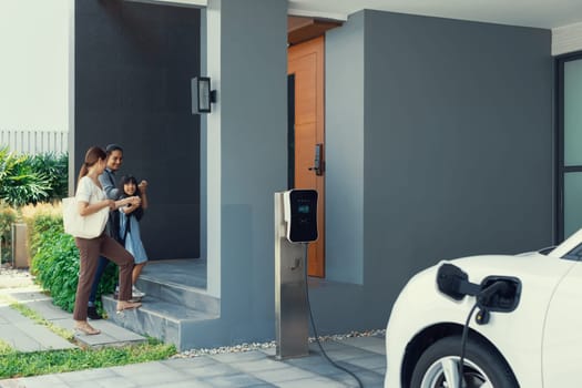 Progressive young parents and daughter with electric vehicle and home charging station. Green and clean energy from electric vehicles for healthy environment. Eco power from renewable source at home.