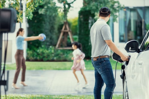 Focus image of progressive man charging electric car from home charging station with blur mother and daughter playing together in the background.