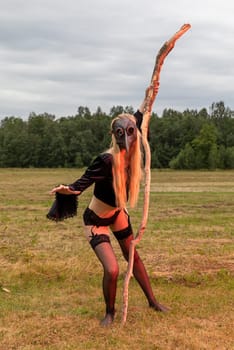 A young woman wearing a black raven mask poses with a dry tree branch in a serene countryside field.