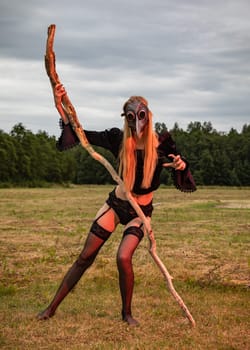 A young woman dressed in black and wearing a black raven mask holds a large dry tree branch and poses in the middle of a field in the countryside