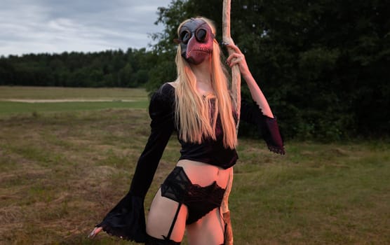 A mysterious woman wears a black raven mask and stands in a field holding a dry tree branch, surrounded by the beauty of the countryside.