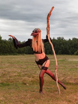 A young woman wearing a black raven mask poses with a dry tree branch in a serene countryside field.