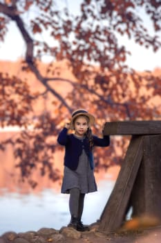 Autumn vintage portrait of a cute girl stand in a straw boater hat, a gray dress and a knitted blouse on a background of red foliage and a pond. Vertical. Copy space