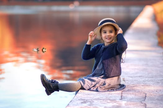 A cheerful little girl sits on the shore of a pond, smiling in vintage clothes, a straw hat, a gray dress and a knitted blouse against a background of a park and red foliage in autumn. Copy space