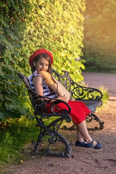 A beautiful brunette little girl in a red beret and a striped T-shirt sits on a bench in the park, holding a bag with French baguettes, smiling at the camera. Copy space