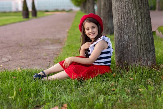 Portrait of a beautiful little brunette girl, dressed in a French style: in a red beret, a striped T-shirt and a red skirt, sits under a tree in a summer park, smiles, looks to the side