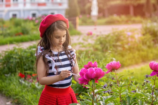 A portrait of a beautiful brunette little girl in a red beret and a striped T-shirt stands next to a bush of peonies in a park in summer, paints them with a brush. Copy space