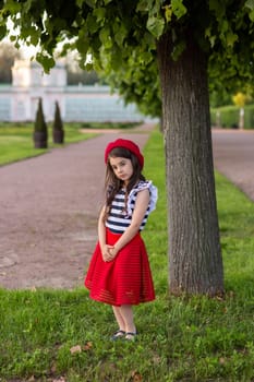 A beautiful little brunette girl, dressed in a French style: in a red beret, a striped T-shirt and a red skirt, stands under a tree in a summer park, looks at the camera