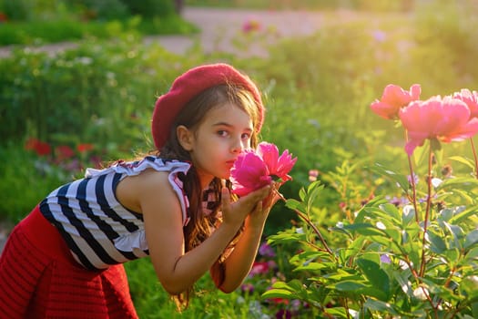 A beautiful brunette little girl in a red beret and a striped T-shirt is holding a large pink peony in the park in summer, sniffing it. Copy space
