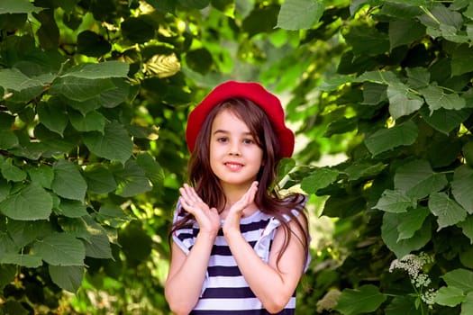 Portrait of a beautiful brunette girl in a red beret and a striped T-shirt stands against a background of green leaves in the park. Copy space