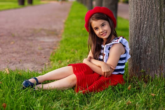Portrait of a beautiful little brunette girl, dressed in a French style: in a red beret, a striped T-shirt and a red skirt, sits under a tree in a summer park, smiles, looks at to camera