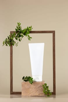 White cosmetic tube, stands on a rectangular beige stone, in an empty wooden frame with branches of a green plant, on beige background . Mock up