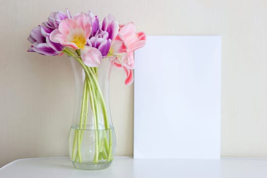A bouquet of open pink and purple tulips in a glass vase stands on a white table against a light wall. Next to it is a white sheet of paper. Vertical