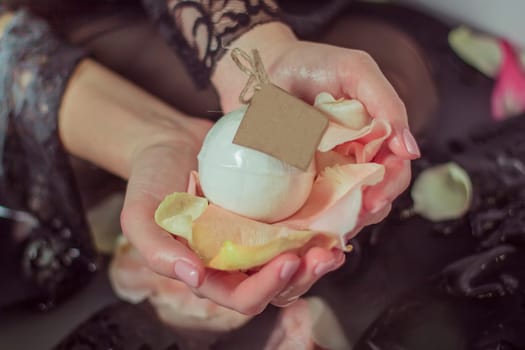 Woman's hands in black lace clothes, holding a ball of salt, above water with an empty label and with pink rose petals. Close up. Mock up