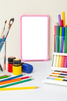 Pink digital tablet on a white table with drawing supplies: brushes, watercolors,crayons, markers, pencil , acrylic paints. The concept of children's drawing courses. Copy space. Vertical