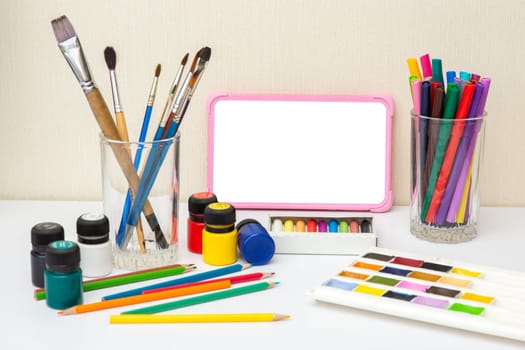 Children's digital tablet on a white table with drawing supplies: brushes, watercolors,crayons, markers, pencil , acrylic paints. The concept of children's drawing courses. Copy space.
