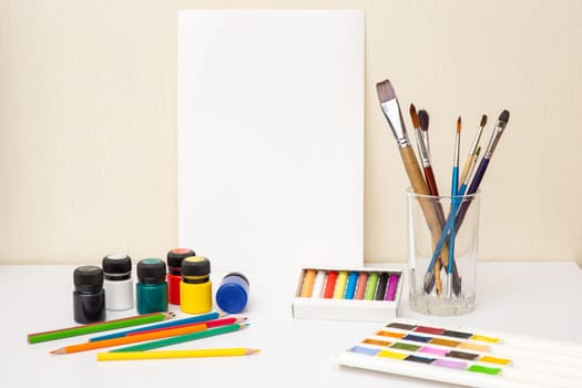Colorful drawing supplies and white paper on white table: Brushes, watercolors,crayons, pencil , acrylic paints. Mock up