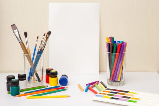 White table with colorful drawing supplies and white paper . Brushes, watercolors,crayons, pencil , acrylic paints. The concept of drawing courses. Mock up