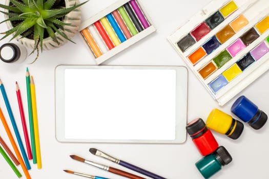 White digital tablet with a blank screen on a white table with drawing supplies: watercolors, pastel crayons, pencil, acrylic paints and succulent pot . Copy space. Mock up