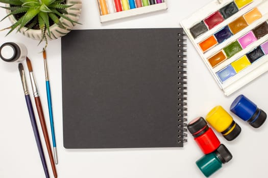 Black square sketchbook on a white table with drawing supplies: watercolors, pastel crayons, brushes, acrylic paints, succulent pot.Copy space.Mock up