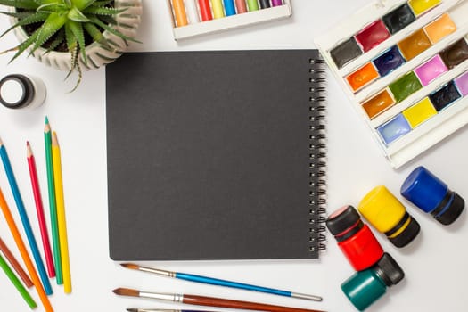 Black sketchbook on a white table with drawing supplies: watercolors, pastel crayons, colored markers, brushes, acrylic paints, succulent pot.Copy space.Mock up