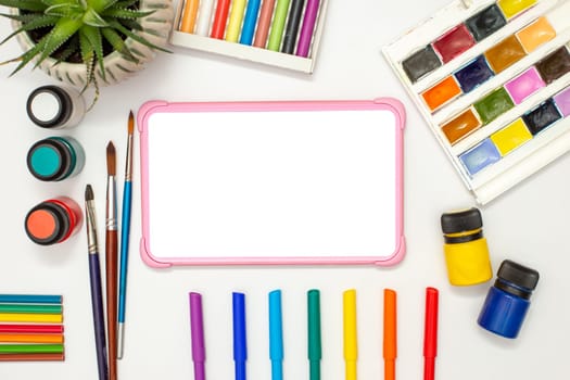 Children's pink digital tablet with a blank screen on a white table with drawing supplies: watercolors, pastel crayons, colored markers, acrylic paints. A succulent pot is standing nearby. Copy space.