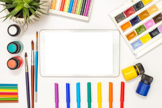 Top view: white digital tablet with blank screen on white table and drawing supplies: watercolors, pastel crayons, colored markers, brushes, acrylic paints, succulent pot . Copy space. Mock up