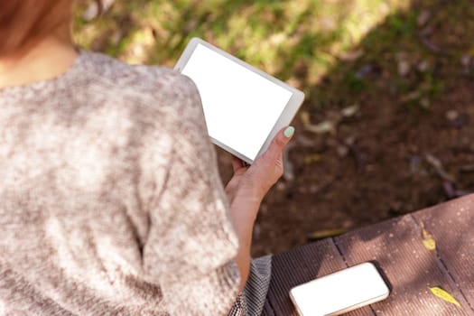 Girl sitting in a knitted jumper in the park holding a digital tablet and smartphone with a white screen. Remote work, education concept. Copy space. No face. Mock up