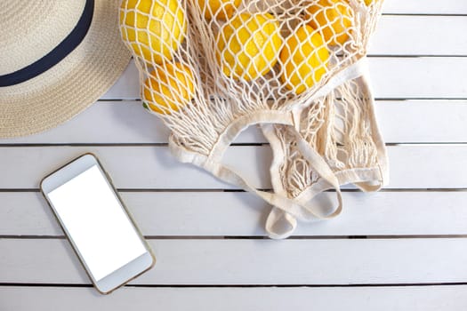 A white smartphone with a white screen lies on a white wooden table, next to it is a white knitted bag with yellow lemons and a straw hat. Mock up. Copy Space