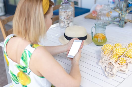 A blonde girl in a summer sundress sits at a white table, holds a white smartphone in her hands, next to it is a jug of lemonade and yellow lemons in a knitted bag. Back view.Mock up