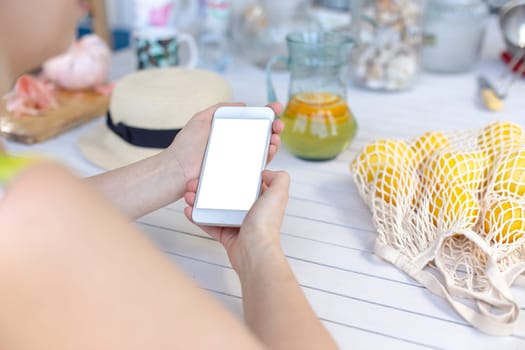 Hands of a girl holding a white smartphone, on a white wooden table, next to yellow lemons in a knitted bag. View from above. Mock up.