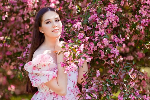 A charming brunette in a pink dress standing near pink blooming apple trees, holding a blooming branch, in the spring in the garden, look at camera, smiling. Copy space