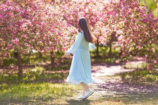 Happy brunette girl in light blue dress, with long hair is dancing in a pink blooming garden, in sunny day. Rear view. Copy space