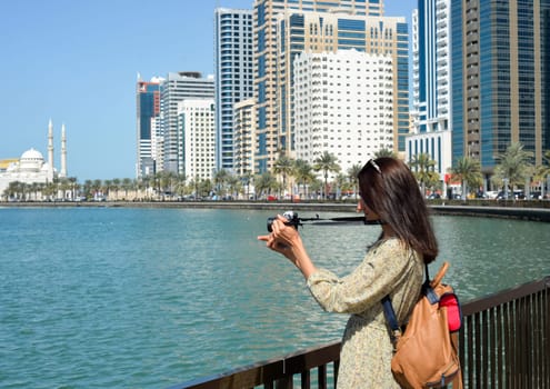 A woman in a long dress with a backpack walks with a camera along the Al Majaz embankment, Lake Khaled, Sharjah emirate. Rear view of a woman photographing the embankment.