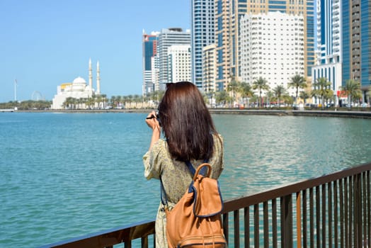 A woman in a long dress with a backpack walks with a camera along the Al Majaz embankment, Lake Khaled, Sharjah emirate. Rear view of a woman photographing the embankment.