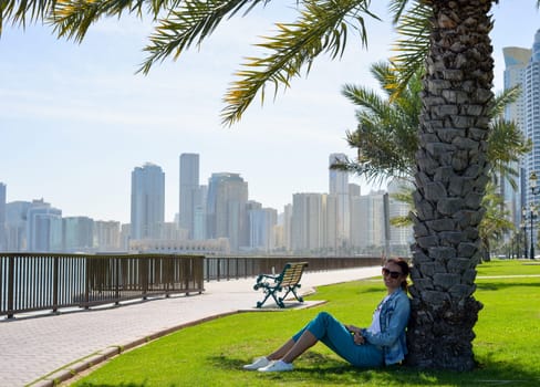 Happy tourist relaxing on lawn under palm tree in Sharjah while looking at camera. Vacation and sightseeing concept.