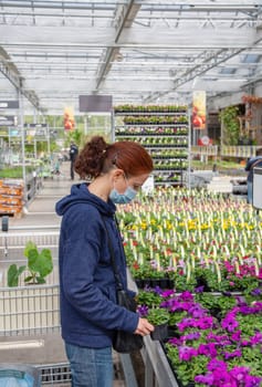 A young woman in a mask chooses and buys seedlings in a garden center with a large assortment of plants, a woman enjoys hobbies and leisure activities, growing plants and flowers in her garden