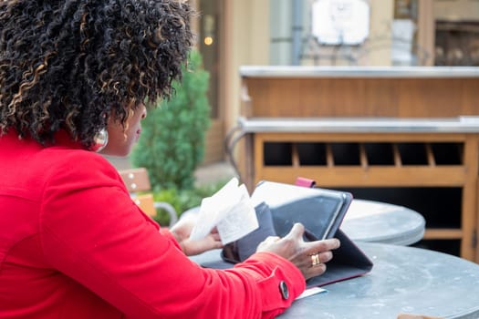 a young african american woman in a red coat sits at a table in a cafe and checking bill after shopping using a tablet, financial problems crisis, proper planning of expenses, High quality photo