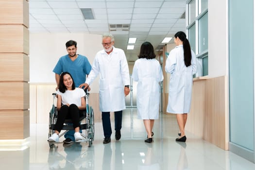 Doctor and male nurse transport a female patient in a wheelchair along sterile hospital corridor. Health care and nursing care for disabled handicapped patient in the hospital concept.