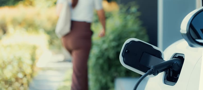 Focus EV charger plugged into EV car at home charging station with blurred background of progressive woman walking in background. Elective vehicle powered by clean energy for eco-friendly concept.