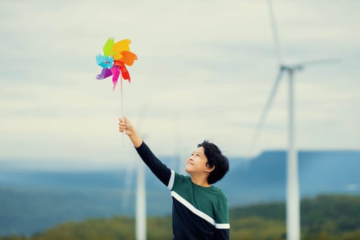 Progressive young asian boy playing with wind pinwheel toy in the wind turbine farm, green field over the hill. Green energy from renewable electric wind generator. Windmill in the countryside concept