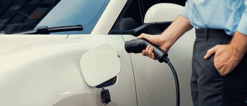 Closeup progressive man holding EV charger plug from public charging station for electric vehicle with background of residential building as concept eco-friendly sustainability energy car concept.