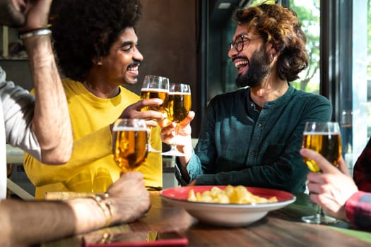 Vertical portrait of happy multiracial male friends in a bar looking at camera toasting with beer. Lifestyle concept.