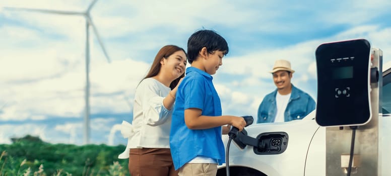 Concept of progressive happy family enjoying their time at wind farm with electric vehicle. Electric vehicle driven by clean renewable energy from wind turbine generator for charging station.