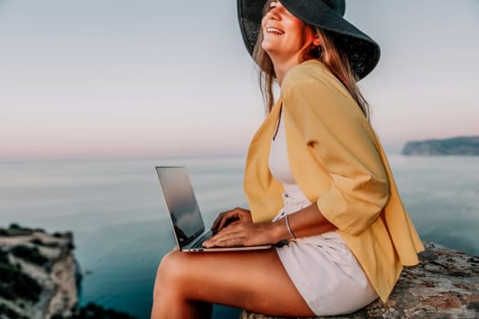 Successful business woman in yellow hat working on laptop by the sea. Pretty lady typing on computer at summer day outdoors. Freelance, travel and holidays concept.