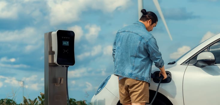 Progressive man with his electric car, EV car recharging energy from charging station on green field with wind turbine as concept of future sustainable energy. Electric vehicle with energy generator.